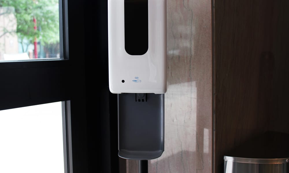 A white hand sanitizer station standing in front of a brown wall at the entrance of an upscale hotel chain.
