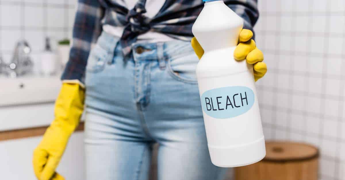A woman holds a bottle with a bleach label. She's wearing yellow gloves and looks like she's going to clean her bathroom.