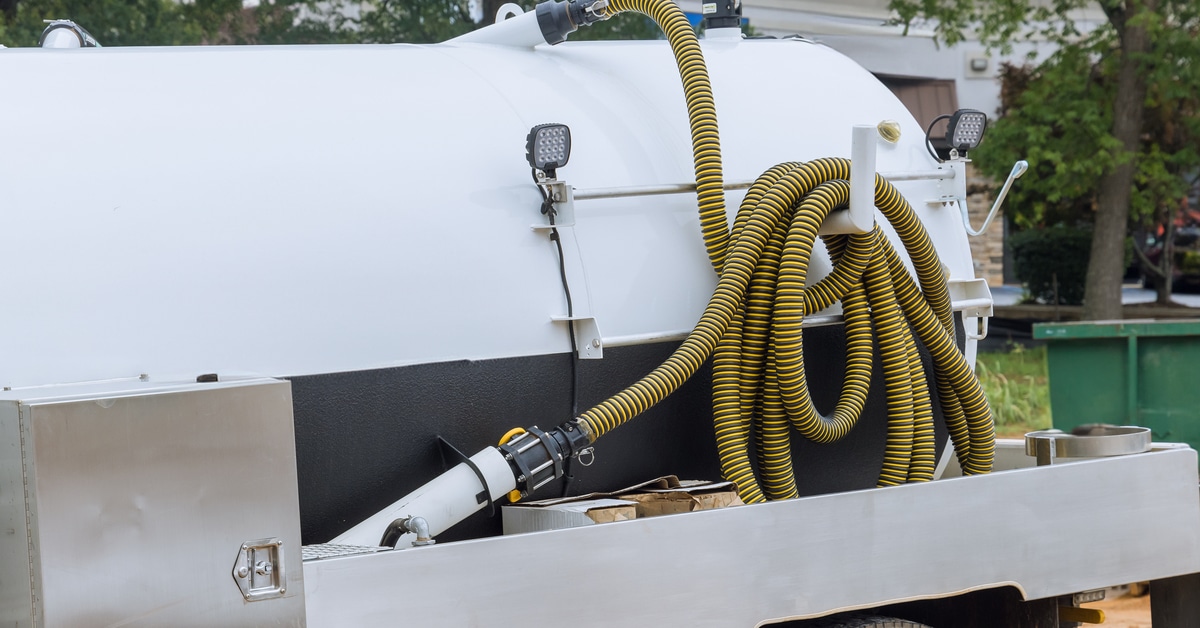 A white septic truck with a yellow and black hose parked near a portable restroom holding tank at a construction site.