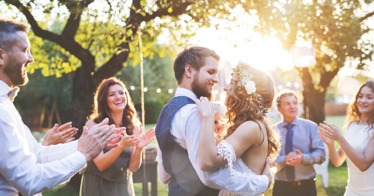 A bride and groom dancing together at an outdoor wedding reception. Friends and family surround them under the setting sun.