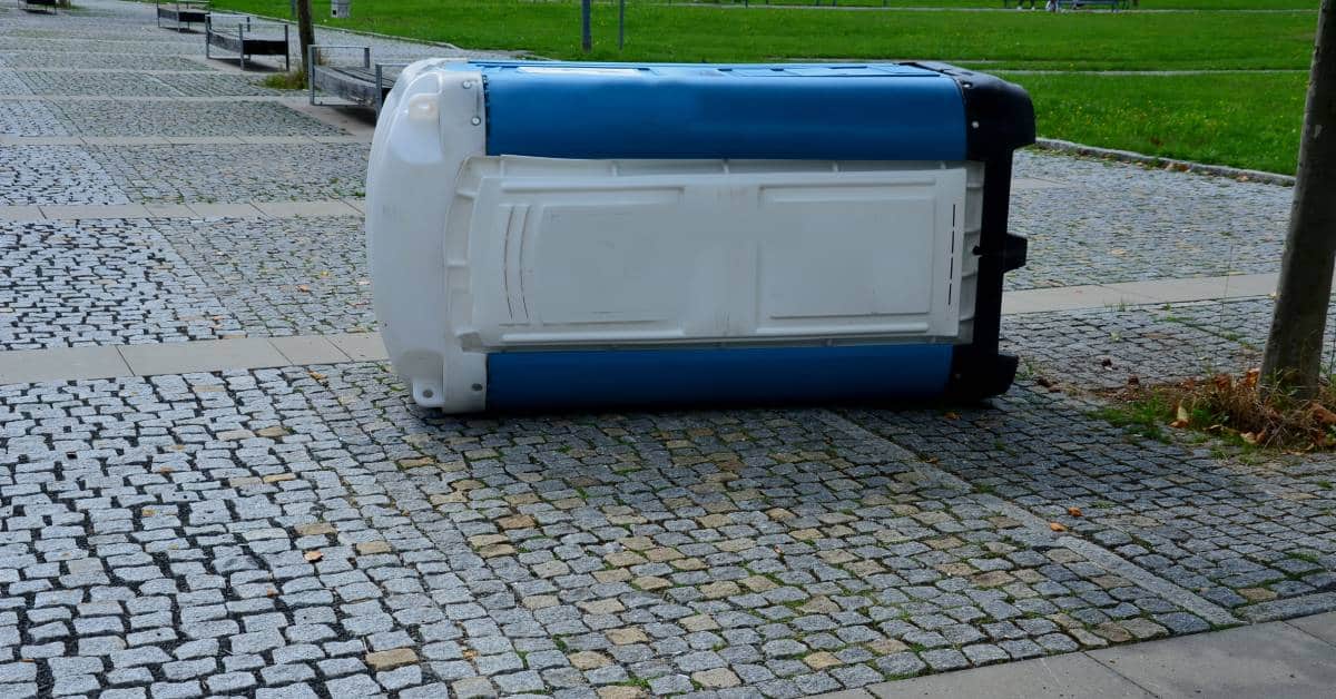 A blue and white plastic porta potty lies on its side in a public park. It rests on a stone walkway.