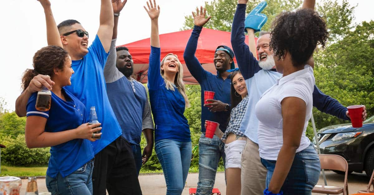 A group of eight individuals is celebrating together at a tailgate party. They raise their hands and smile at each other.