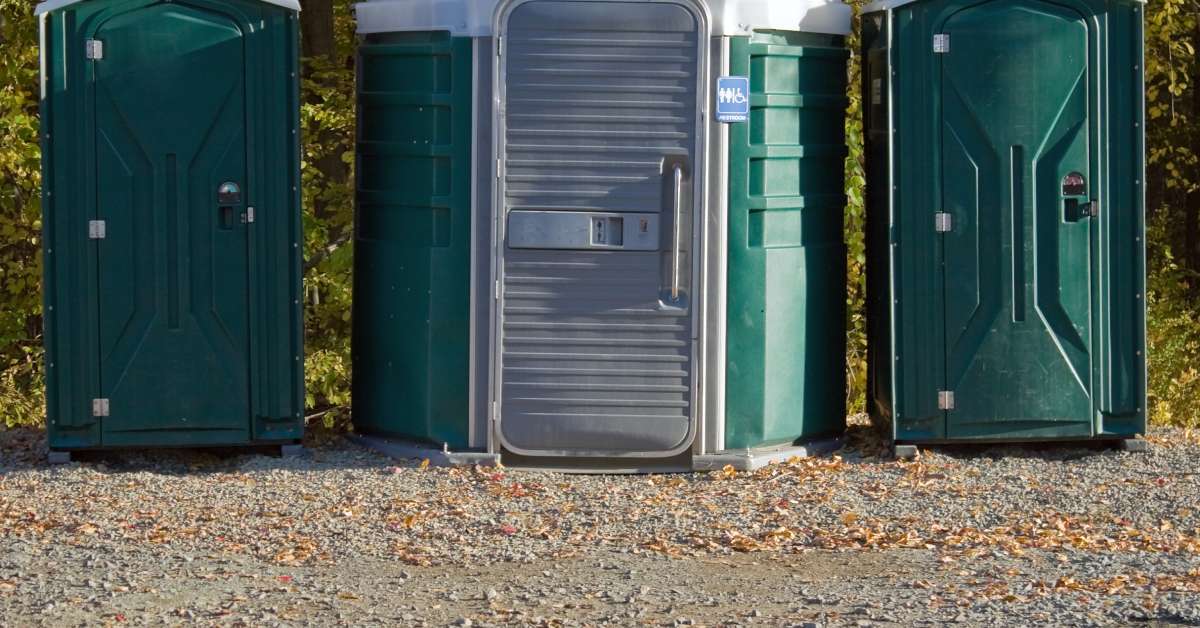 Three porta potties stand outside on a gravel road with trees behind them. The one in the middle is wheelchair accessible.