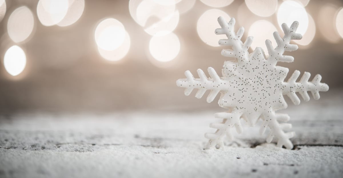 A large white snowflake with colorful glitter resting on a bed of snow. There are bright, blurry lights behind the snow.