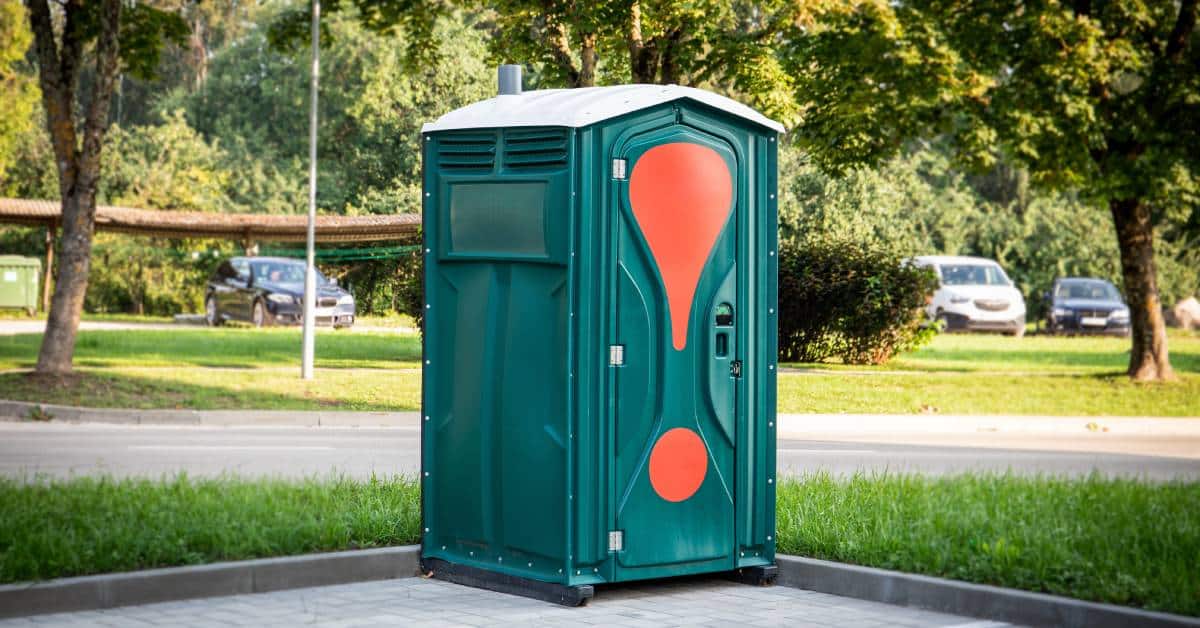 A green porta-potty with orange designs on the door stands outside in a parking lot. Cars are parked in the distance.
