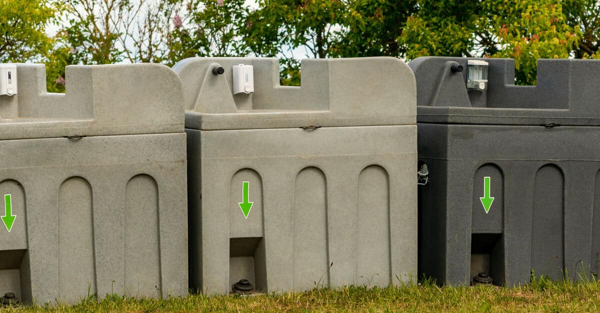 Three gray pedal-powered portable handwashing stations in the grass with lush trees behind them on a cloudy day.