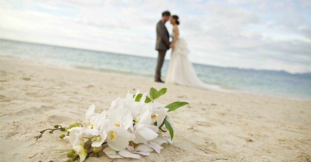 A bouquet of white flowers lying on the sand with a bride and groom standing by a body of water in the background.