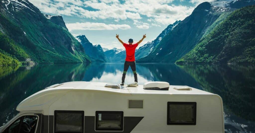 A man raises his arms while standing on top of an RV, overlooking a large lake with mountains surrounding it.
