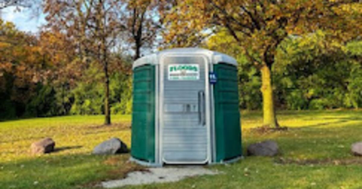 A singular, large portable bathroom placed in the middle of a park. There are large trees just behind it.