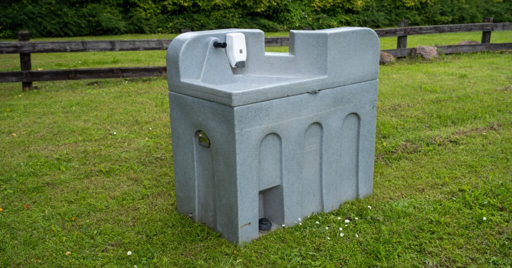 A gray portable hand-washing station sits alone in a grassy field with small white flowers near its base.