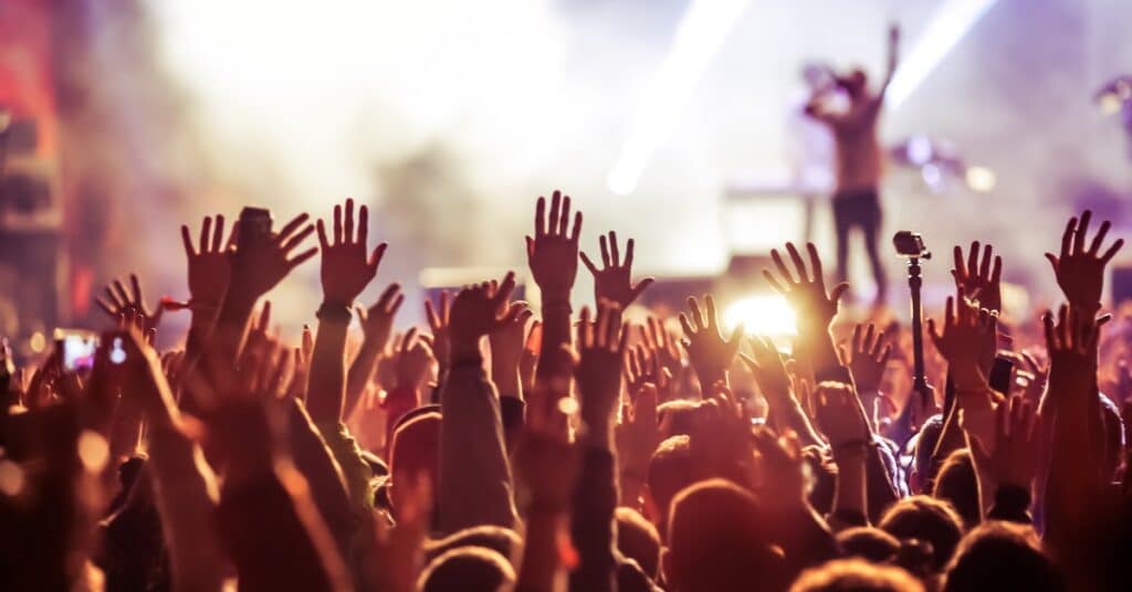 A large crowd of people at a music concert with their hands held in the air. The performer is behind them on stage.