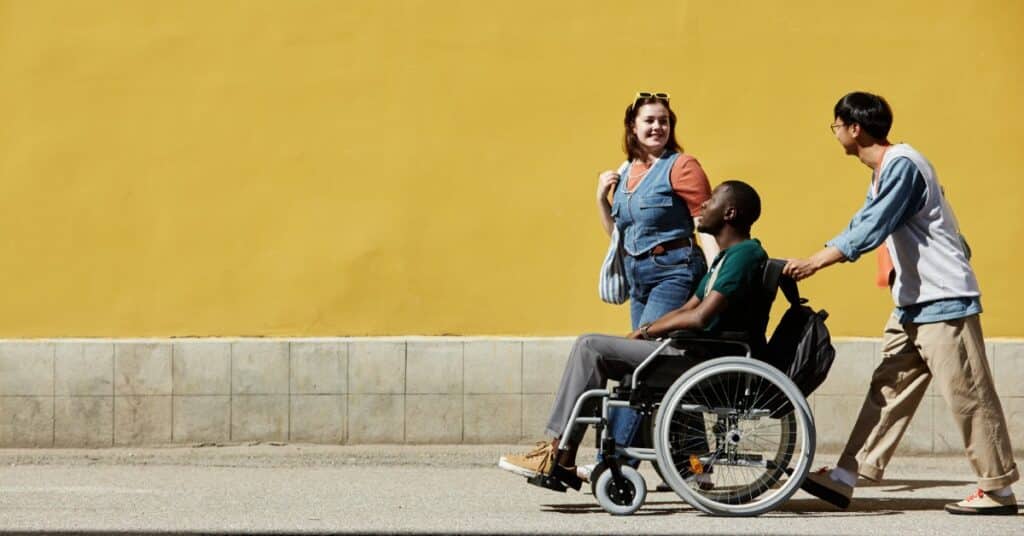 A group of three friends walking down the street. One of them is helping to push another one who is in a wheelchair.