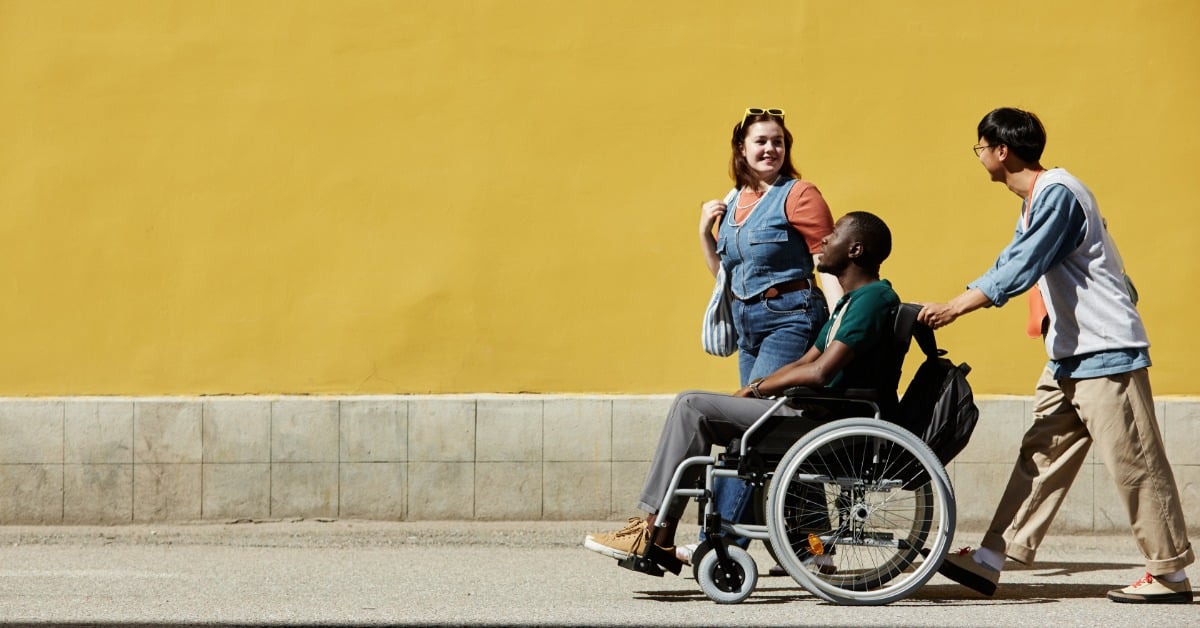 A group of three friends walking down the street. One of them is helping to push another one who is in a wheelchair.