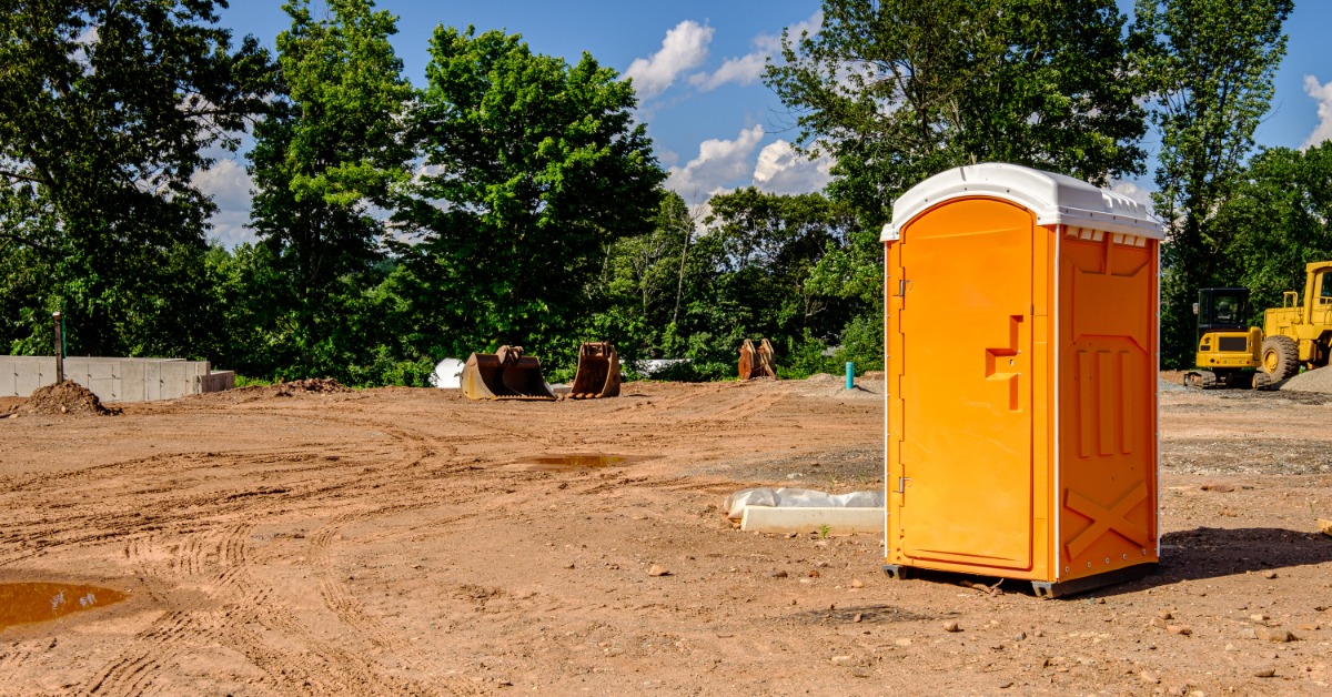 A dusty construction site being readied for work. There is a single orange porta potty placed on the work site.