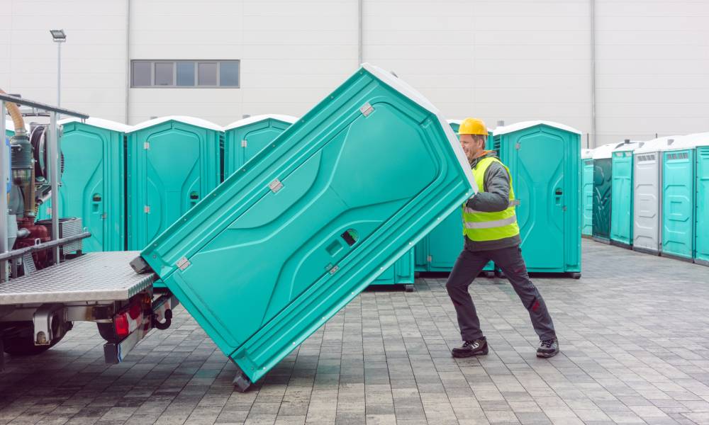 A male worker wearing a yellow hardhat and protective vest loading a light blue rental porta potty onto a delivery truck.
