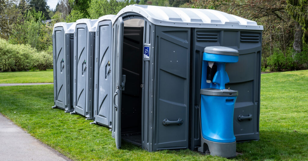Four porta potties are lined up together on a grassy field. There is a handwashing station directly next to the restrooms.
