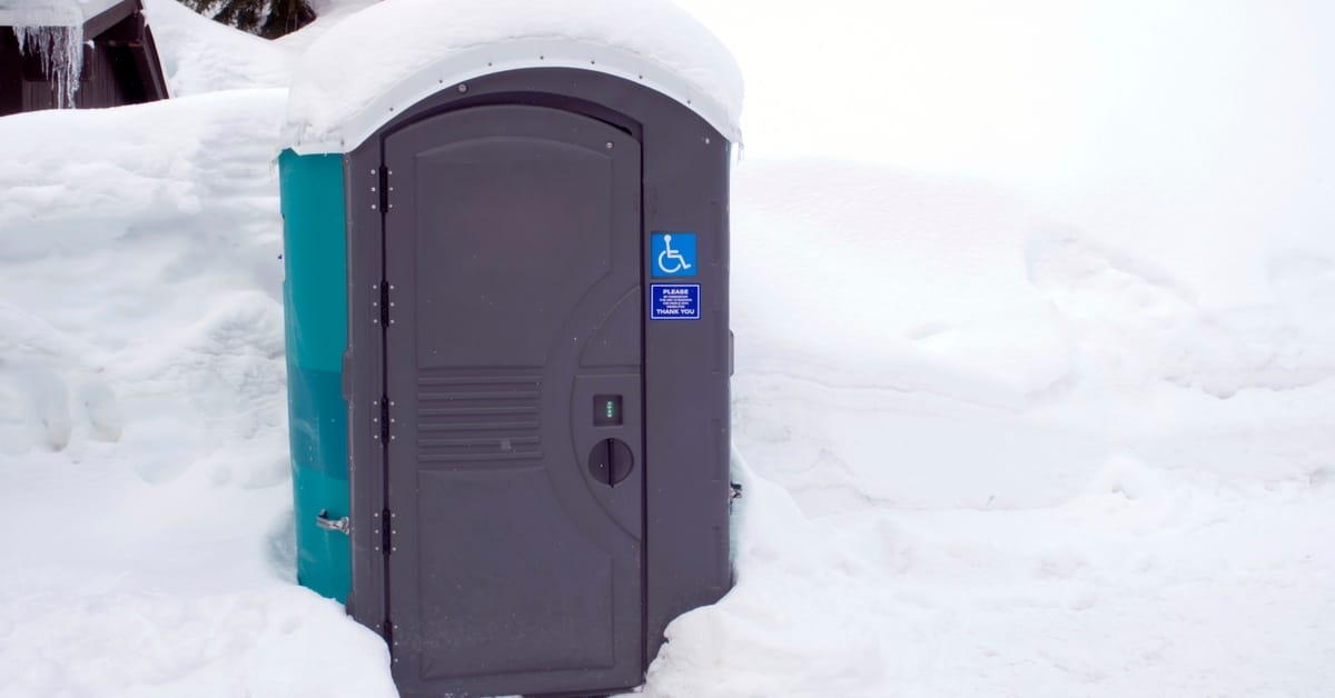 An accessible porta-potty, marked with a handicap symbol sticker, is covered in snow and surrounded by a winter landscape.