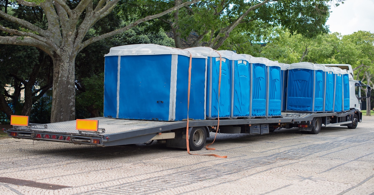 A flatbed truck pulls multiple blue portable toilets, each with orange tie-down straps, down a tree-lined street.