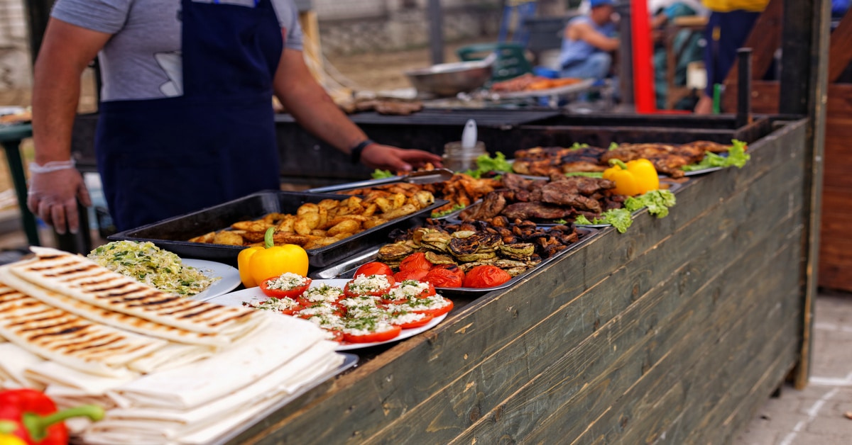 A chef at a food festival stand prepares grilled meats, roasted potatoes, and vegetables, alongside fresh flatbreads.