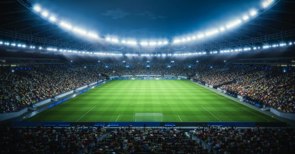 A packed stadium with bright lights illuminates a green soccer field at night, with crowds filling the seats on all sides.