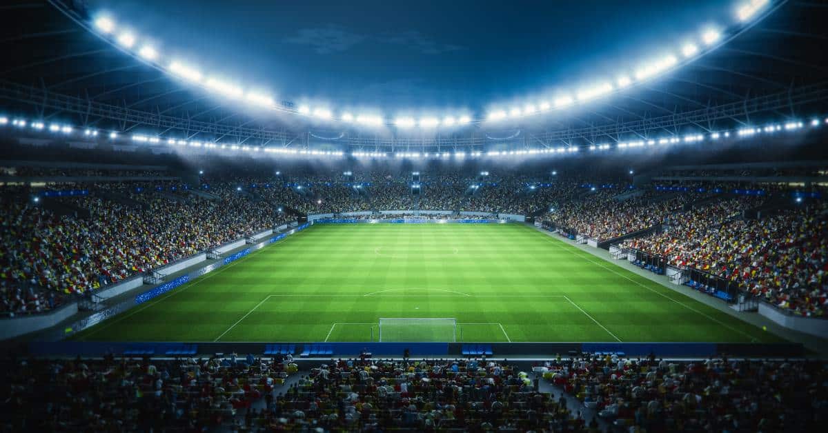 A packed stadium with bright lights illuminates a green soccer field at night, with crowds filling the seats on all sides.
