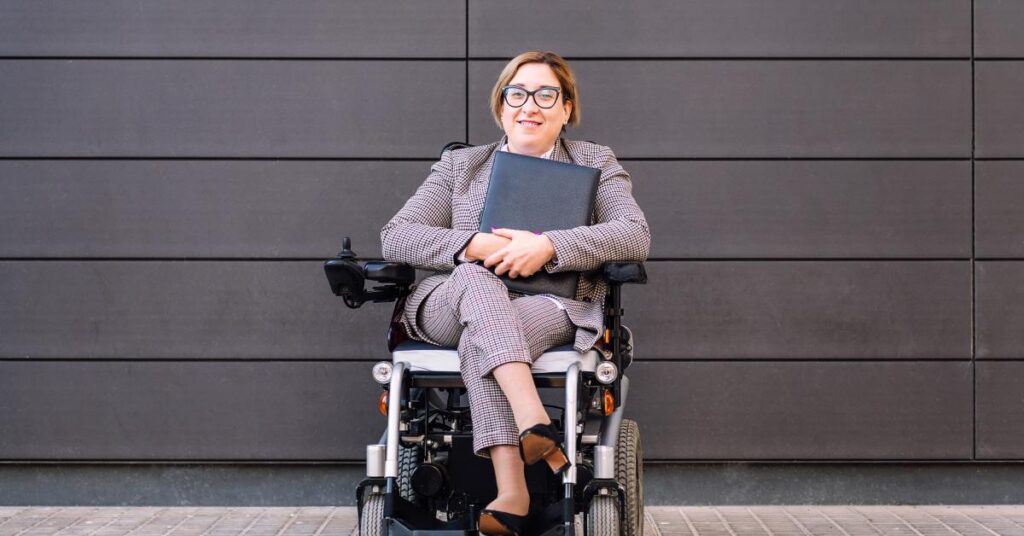 A businesswoman wearing a checkered suit, glasses, and high heels holds a folder and smiles while sitting in a wheelchair.