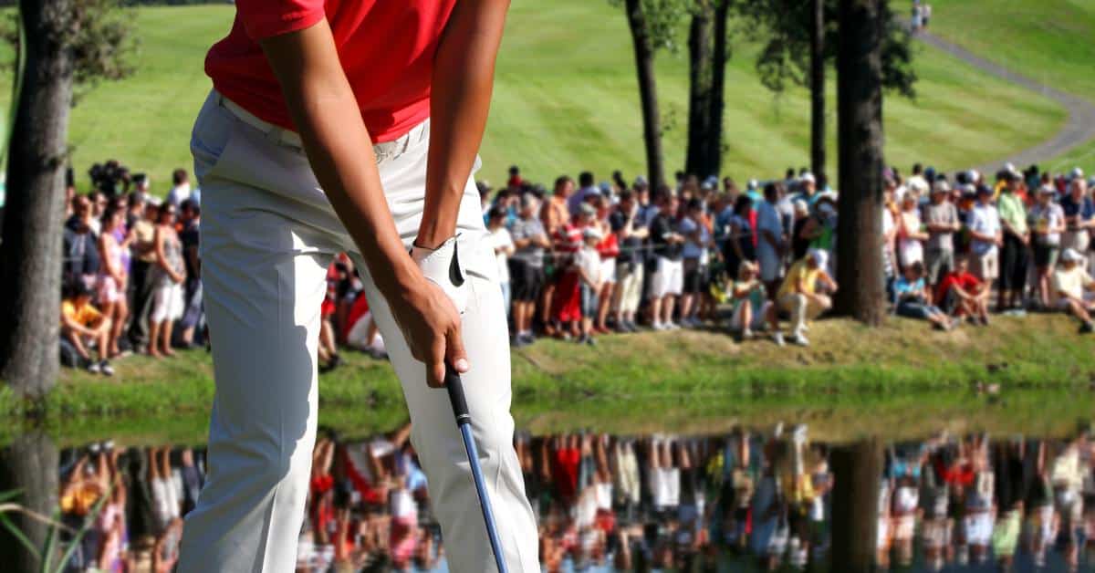 A golfer in a red shirt prepares to putt at a golf tournament as spectators watch from around the green and water hazard.