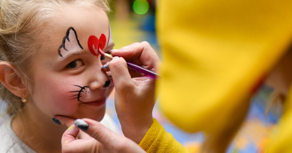 A young girl looks ahead while having her face painted at a children's event full of color and excitement.