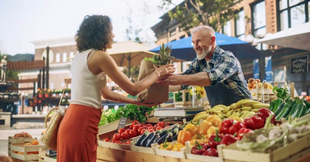 A vendor hands a bag of fresh produce to a customer at an outdoor farmers market with colorful fruits and vegetables on display.