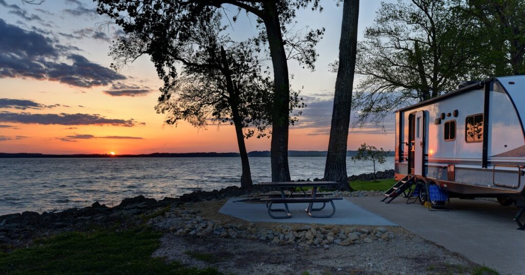 An RV trailer parked at a lakeside campground during sunset, with a picnic table and trees silhouetted against a colorful sky.