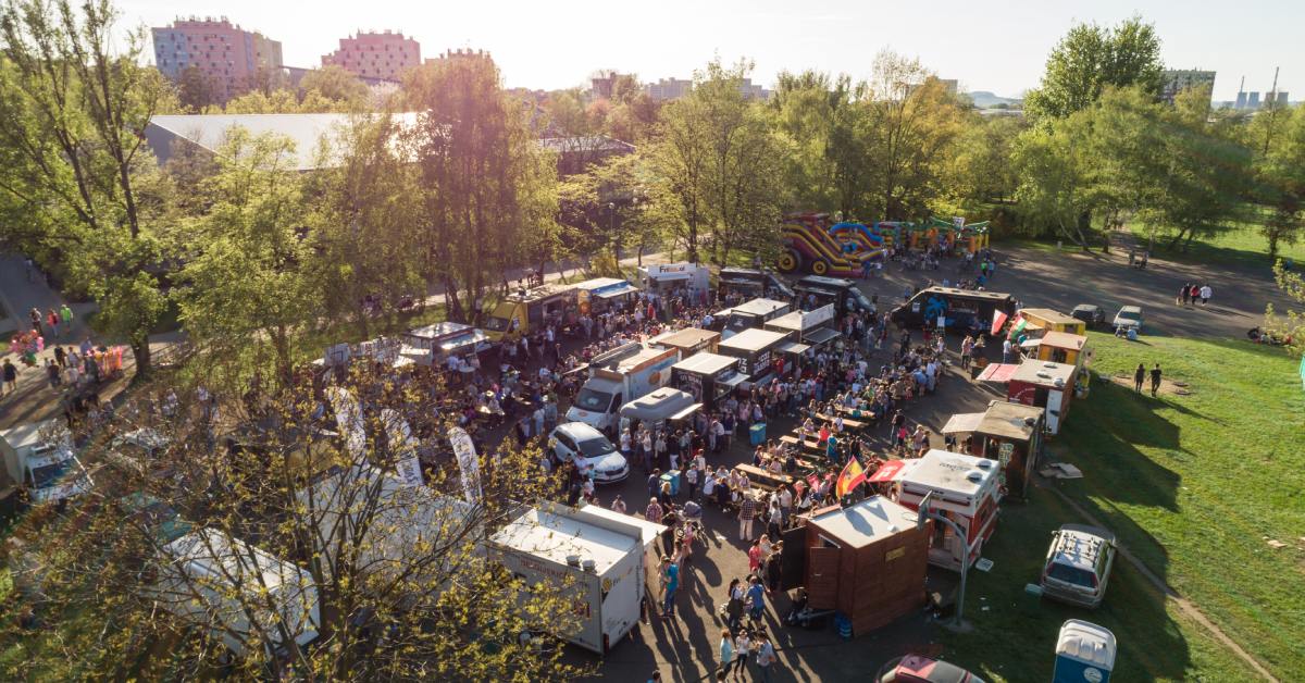 An aerial view of a crowded outdoor food festival with food trucks, picnic tables, and families gathered in a sunny park.