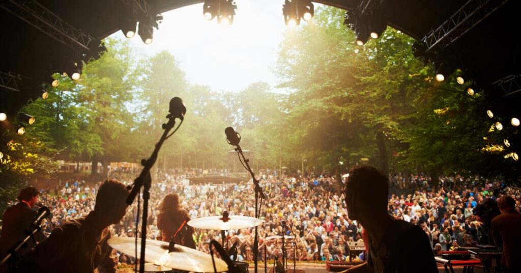 A live band performs on stage at an outdoor concert with a large audience gathered in front during a daytime event.