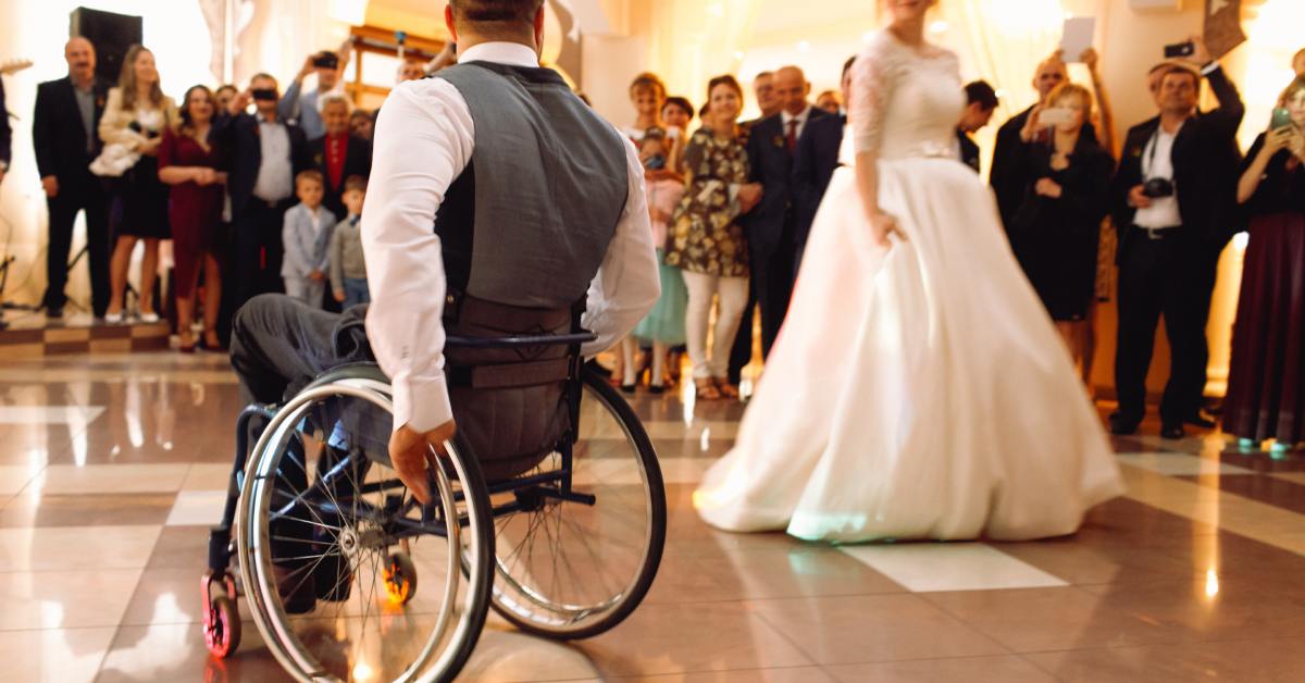 A groom in a wheelchair dances with a bride in a white gown at a wedding reception as guests watch and smile.