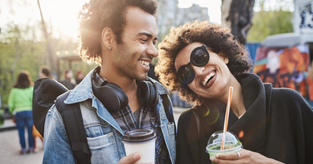 Two friends smiling and holding drinks at an outdoor urban festival, enjoying a lively crowd and sunny city atmosphere.