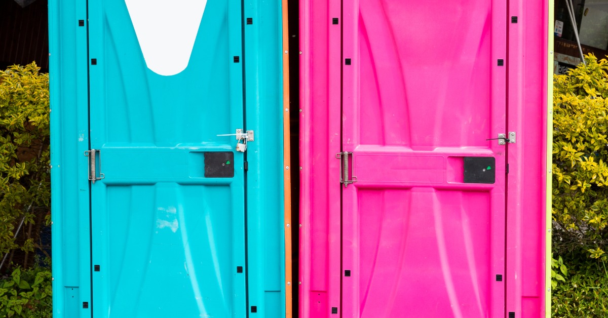 Two colorful portable restrooms side by side—one teal with a white sign, one hot pink—on a brick path outdoors.