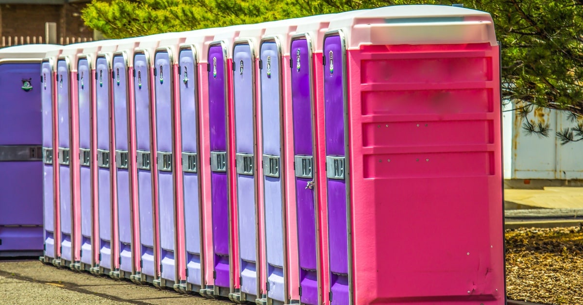 A long row of pink and purple portable restrooms lined up in a paved outdoor area next to green trees.