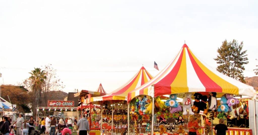 Outdoor carnival game booths with red and yellow striped tents and stuffed animal prizes displayed for fairgoers on a sunny day.
