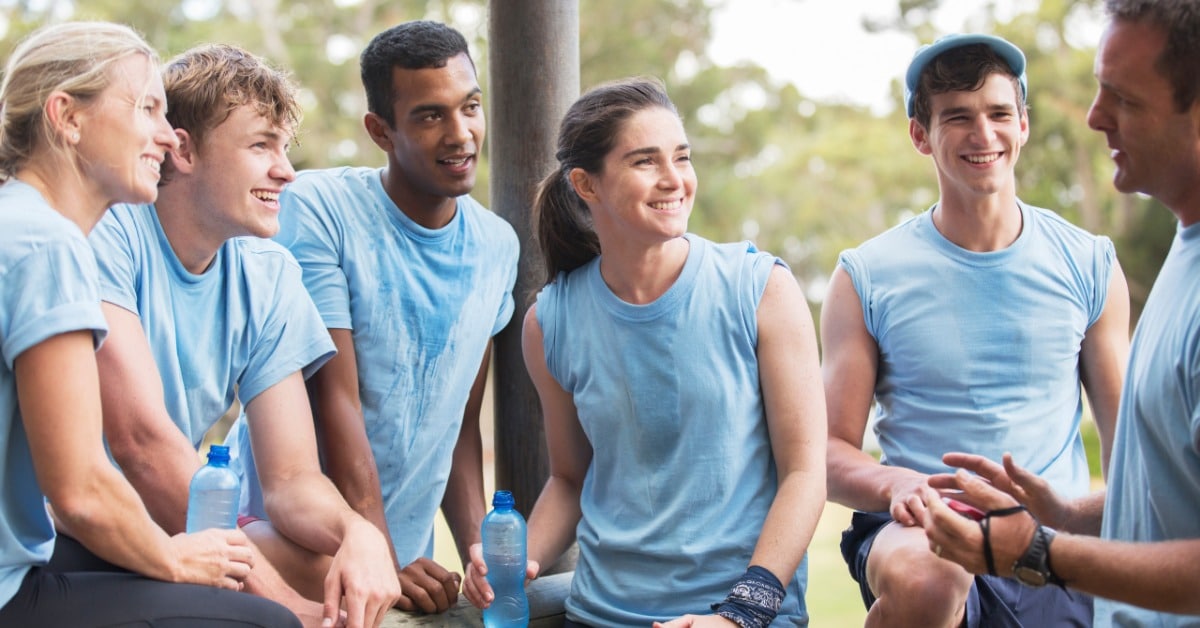 Six teammates in matching blue shirts chat and laugh together at an outdoor team building camping event.