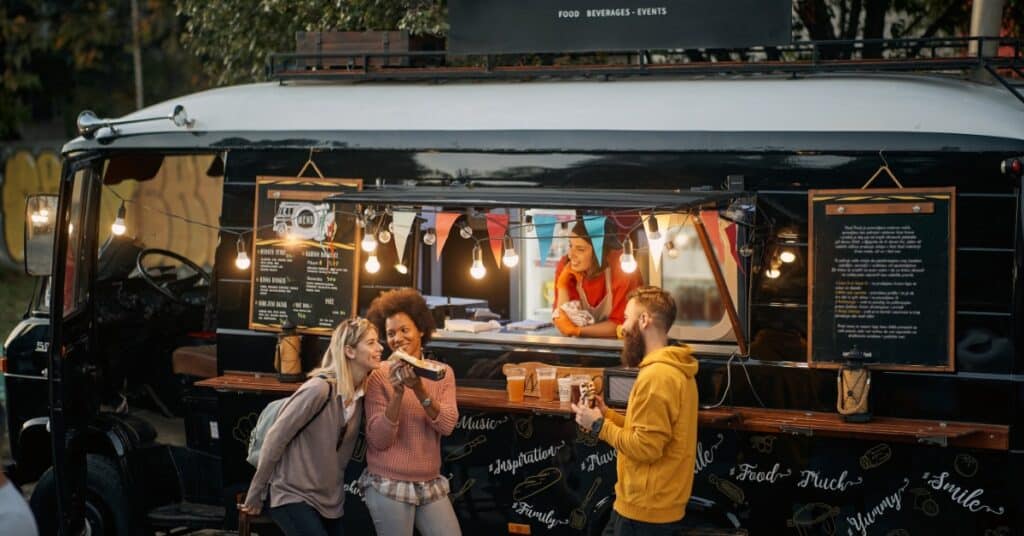 Customers enjoying food and drinks outside a decorated food truck at an outdoor evening event with warm string lighting.
