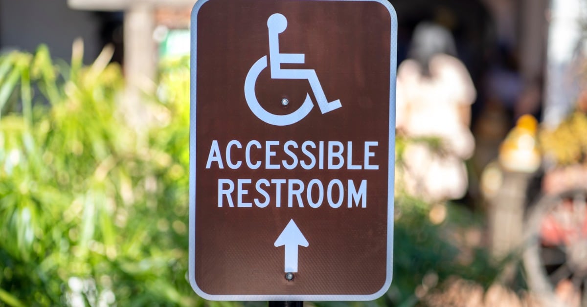 A brown outdoor sign with a wheelchair symbol and upward arrow directing guests toward an accessible restroom facility.