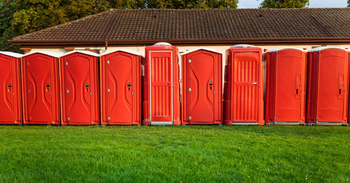 A row of red portable restroom units lined up on green grass in front of a building with a tiled roof.