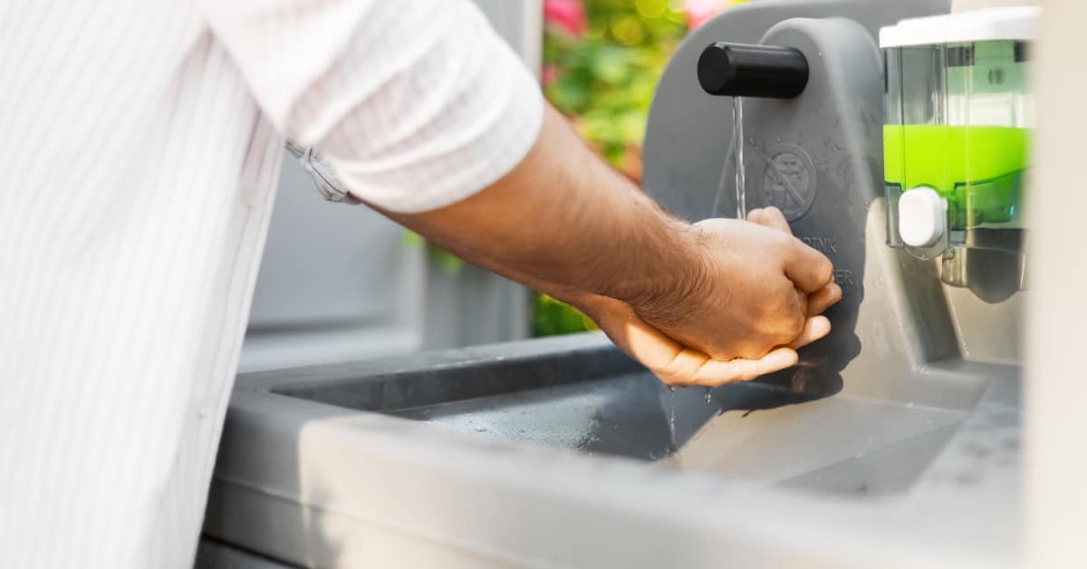 A man washes his hands at an outdoor portable sink with running water and a soap dispenser attached.