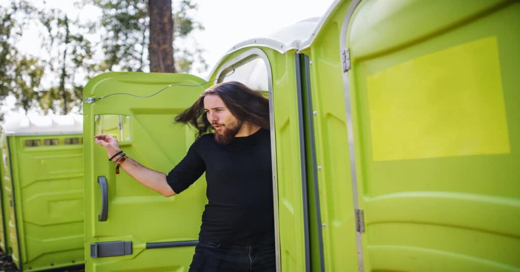 A long-haired, bearded young man in a fitted black top steps out of a bright green portable toilet with a pleased expression.