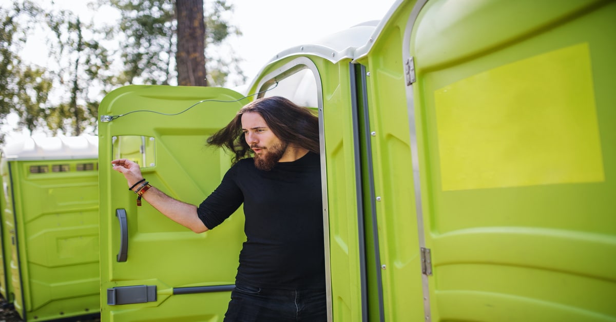 A long-haired, bearded young man in a fitted black top steps out of a bright green portable toilet with a pleased expression.